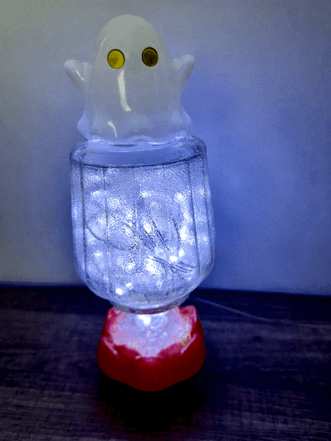 A cute translucent white ghost figurine with yellow eyes sits atop a vintage glass bottle with glowing usb fairy lights