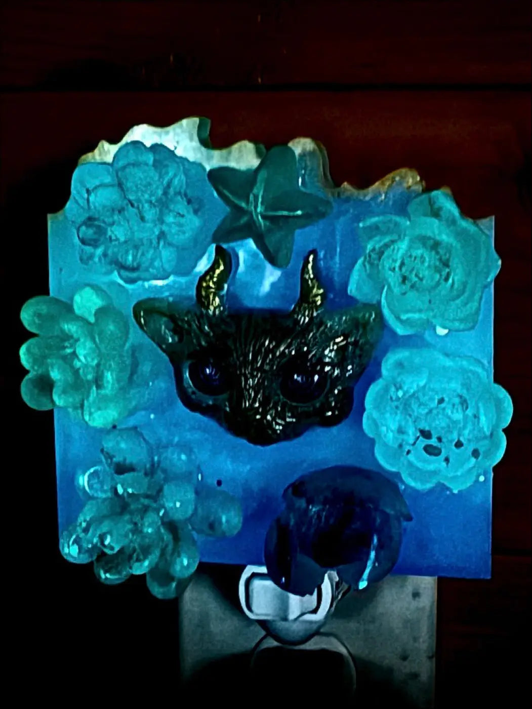 A black glossy horned kitty cat figurine with large eyes sits among teal resin flowers on a garden wall night light base