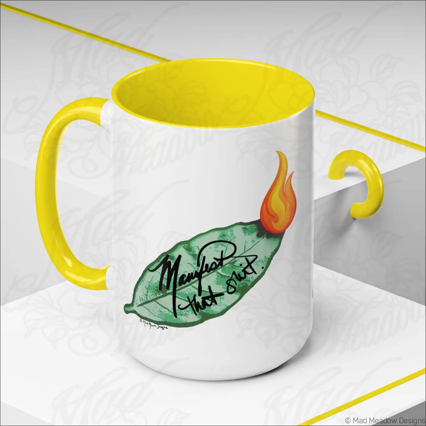 White ceramic coffee mug with glossy finish, bright yellow colored interior, burning bay leaf design, and manifest that shit text