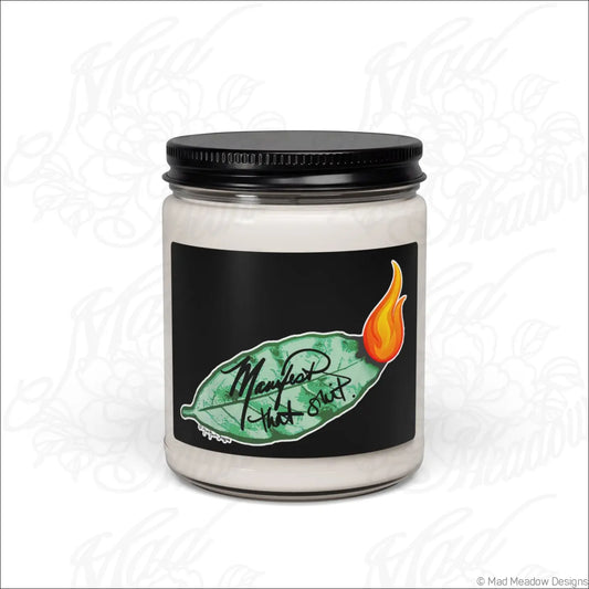 A white soy candle in a clear glass jar with a black lid and bay leaf design labeled manifest that sh*t scented soy wax blend