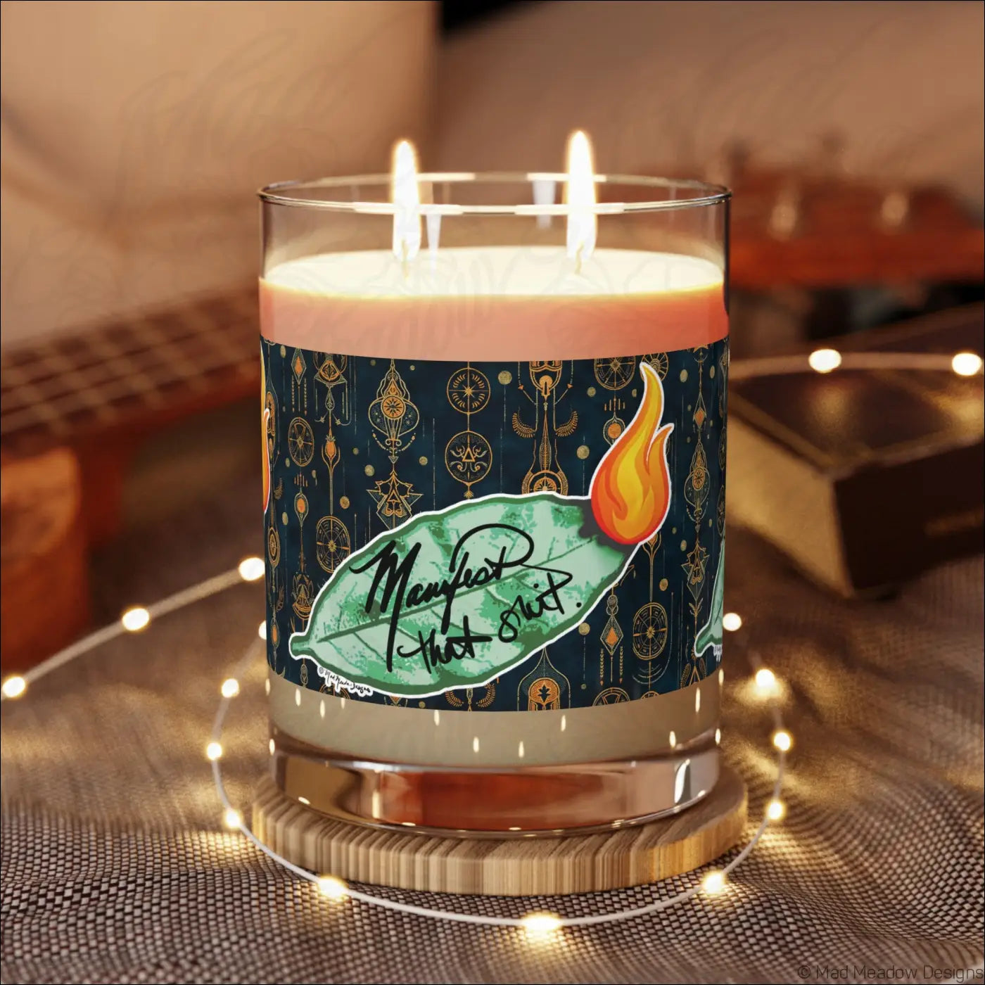 A lit triple-wick glass candle with a dark teal ornate label and bay leaf design for manifestation rituals