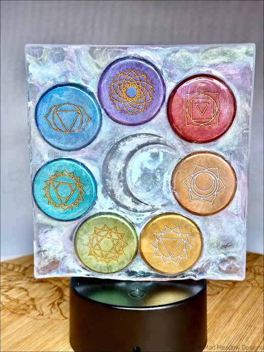 A square resin coaster with seven colorful chakra night light symbols outlined in gold and arranged like rainbow-colored disks