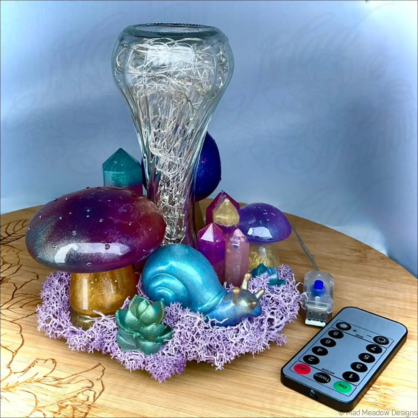 A translucent glass tabletop light with intricate wirework surrounded by colorful handcrafted resin mushrooms crystals and a snail companion on purple moss