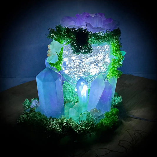 A glowing vintage glass lantern decorated with crystals, flowers, moss, and usb powered fairy lights inside
