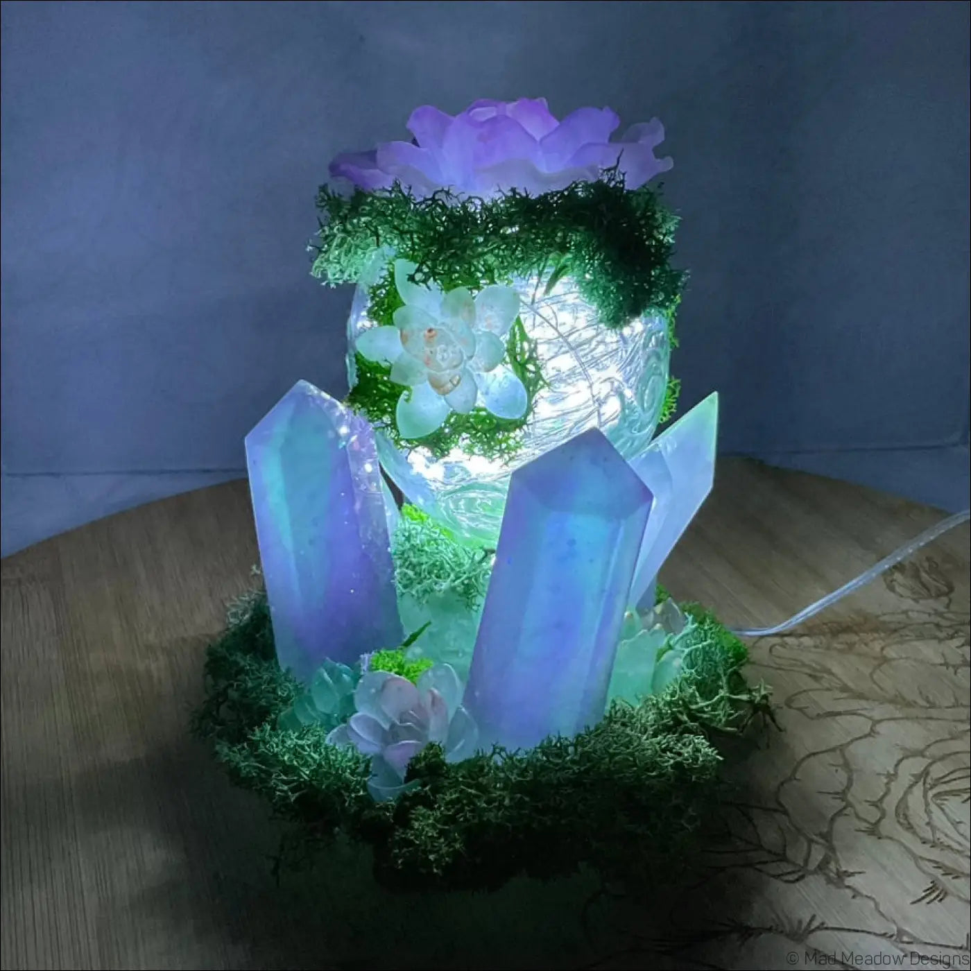 A glowing usb powered vintage glass lantern with green moss, white purple flowers, and blue crystal pillars
