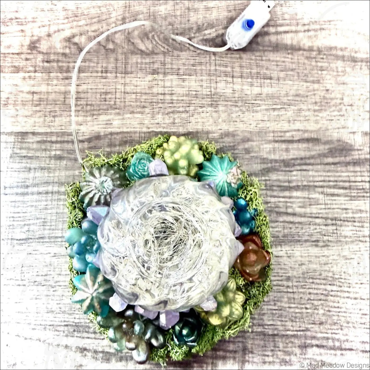 A usb powered crystal filled garden light with a glass bottle and woven wire design surrounded by green and blue succulents