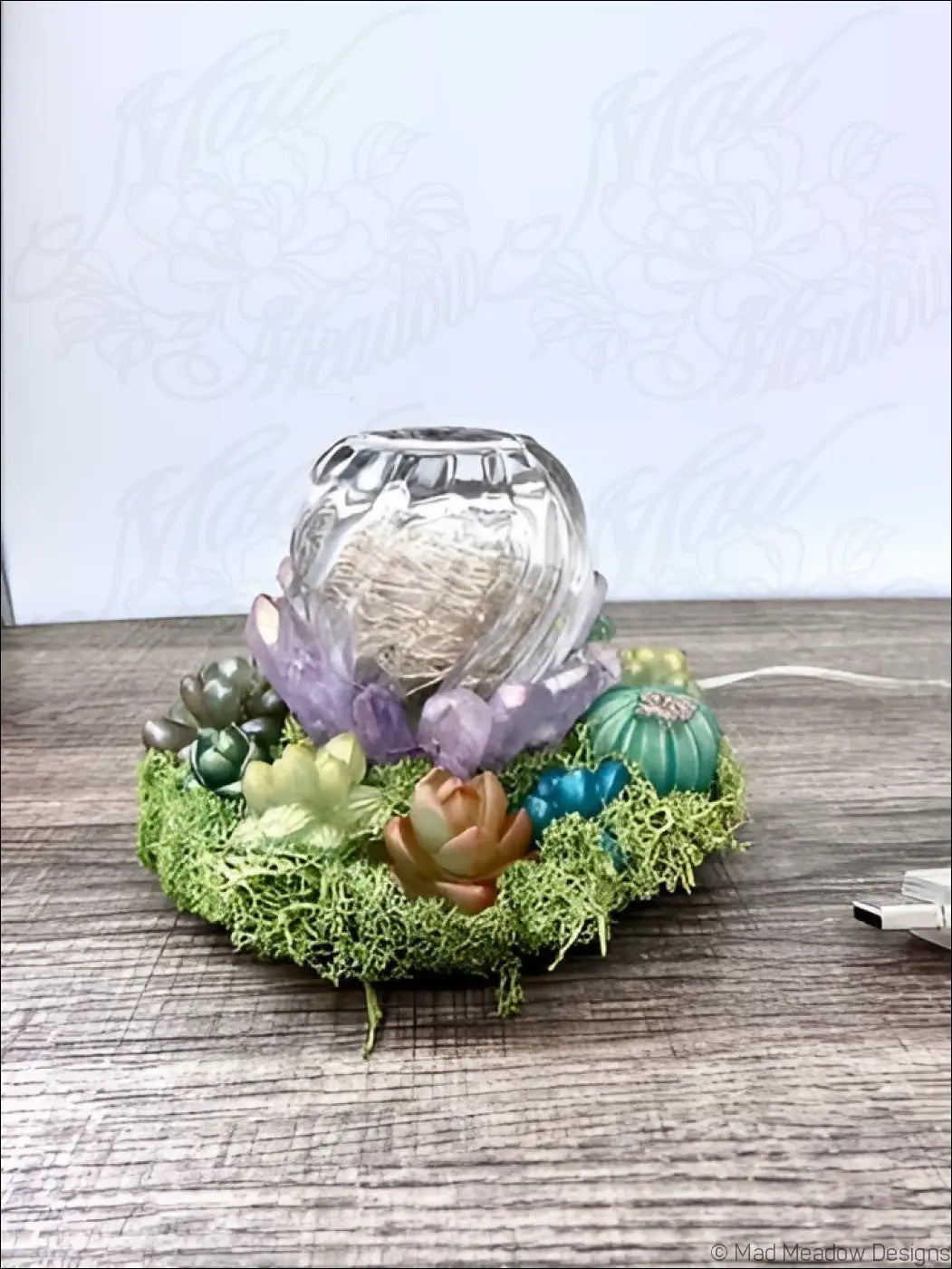 A clear glass usb powered garden light with textured rim sits on green moss surrounded by purple teal and brown succulents