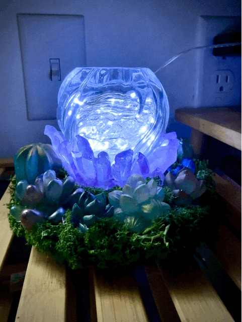 A glowing blue glass orb usb powered light sits on artificial green moss and miniature succulents in a resin garden