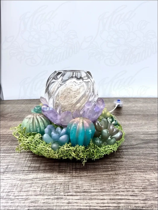 A clear glass usb powered lantern with scalloped rim sits on green moss with purple and teal succulent resin decor