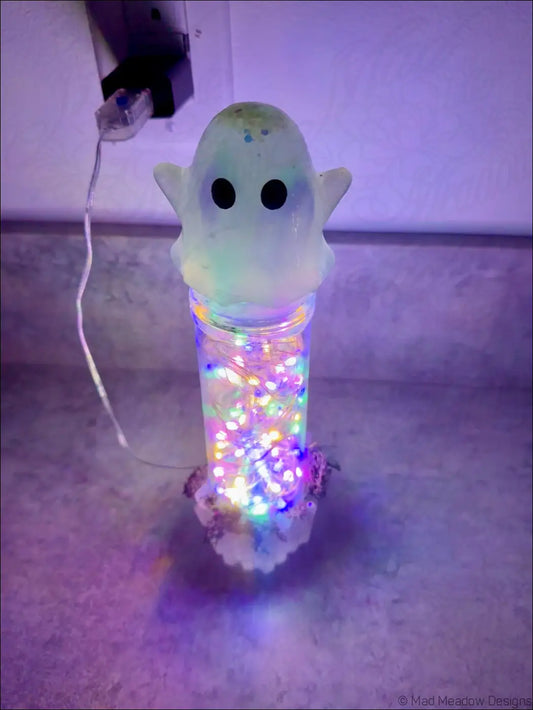 A translucent glowing ghost usb light with black eyes sits on a glass jar filled with colorful led lights and moss