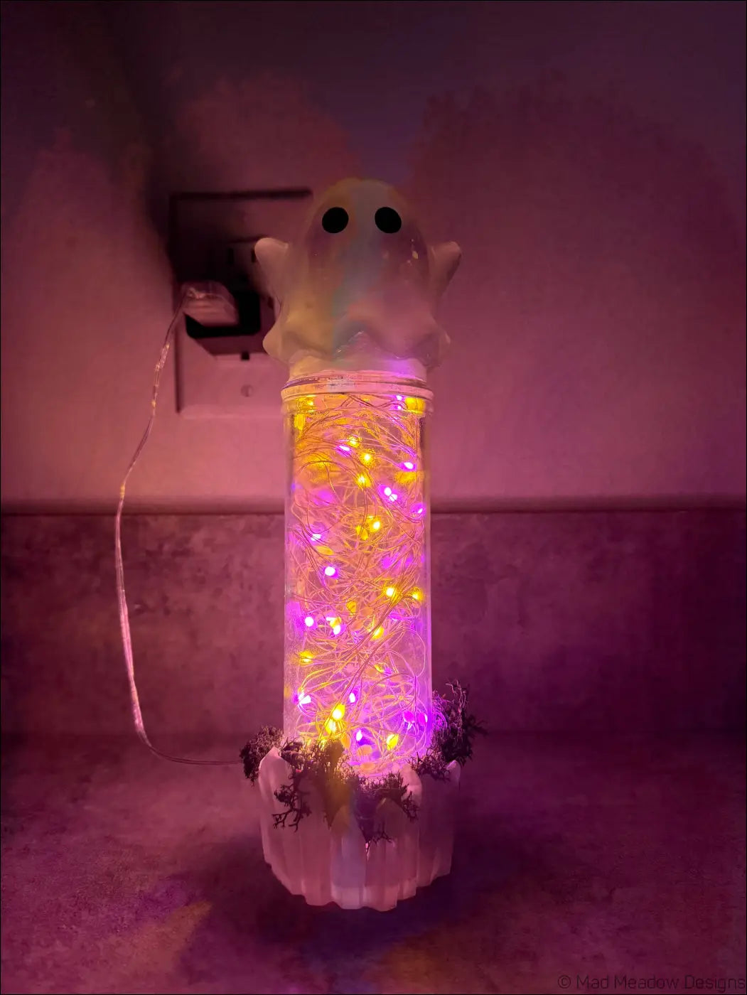 A cute glowing ghost usb light with purple and yellow fairy lights inside a clear cylindrical container with moss and crystals