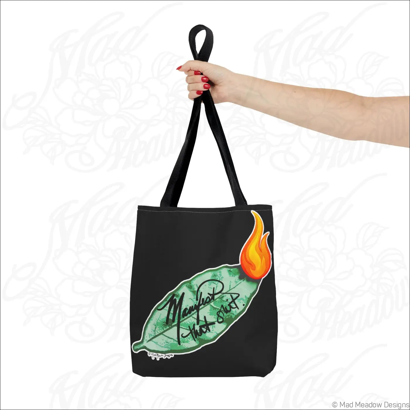 Black canvas tote bag with green bay leaf and orange flame design featuring manifest that in black cursive script with boxed corners and care instructions