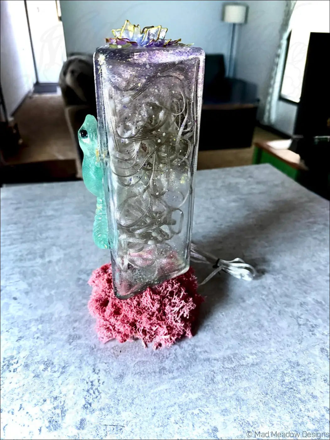 A translucent glass usb lamp with etched designs topped with a gold and purple floral ornament and a turquoise frog figurine