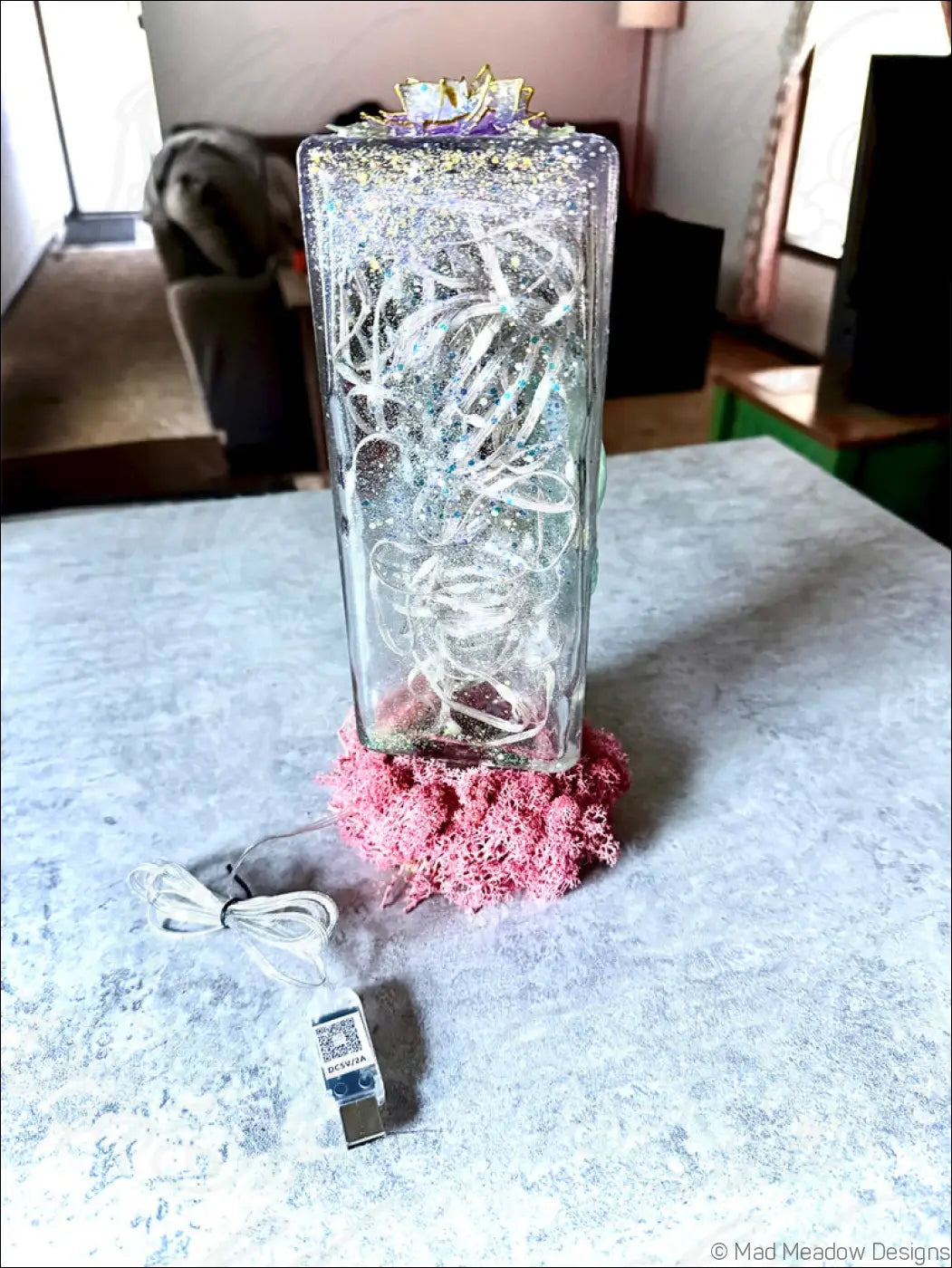 A clear glass rectangular usb lamp with embedded silver wire glitter topped with purple floral design on pink moss