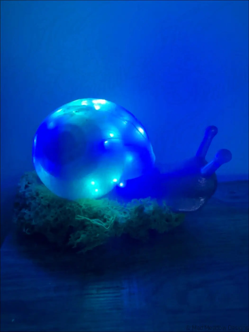 Glowing blue led snail figurine with translucent shell and clear body resting on green moss base with usb light color change