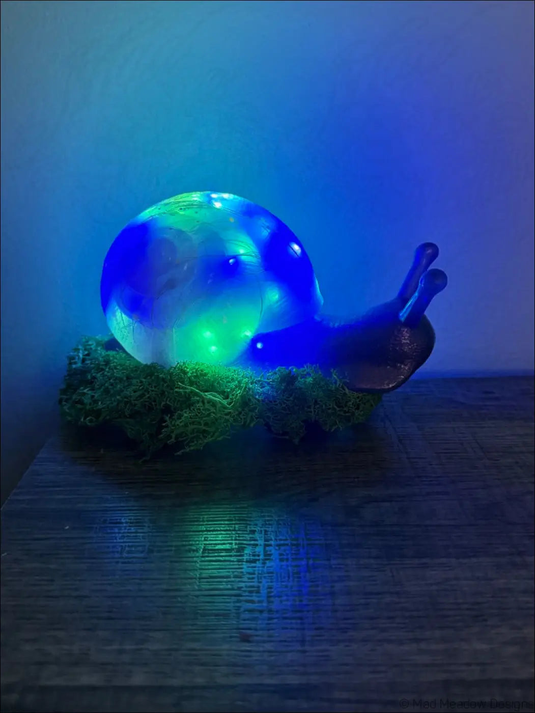 Giant pastel blue and purple snail figurine with glowing translucent shell resting on green moss light usb color change base