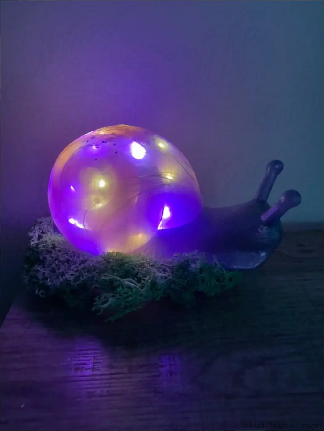 A translucent giant pastel blue and purple snail led lamp with green moss and glowing purple and yellow light inside shell