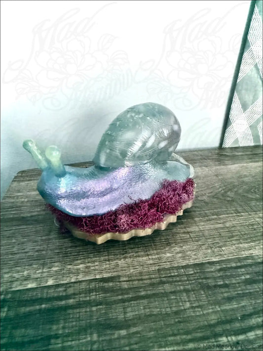 A translucent giant pastel blue and purple snail figurine with a shimmering shell on textured red moss base