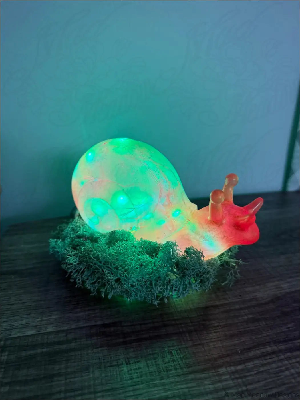A glowing giant pink and gold snail-shaped lamp with green and orange rgb light and red antennae on moss