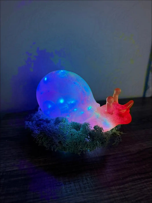 A glowing giant pink and gold snail-shaped led lamp with a translucent blue shell resting on artificial moss