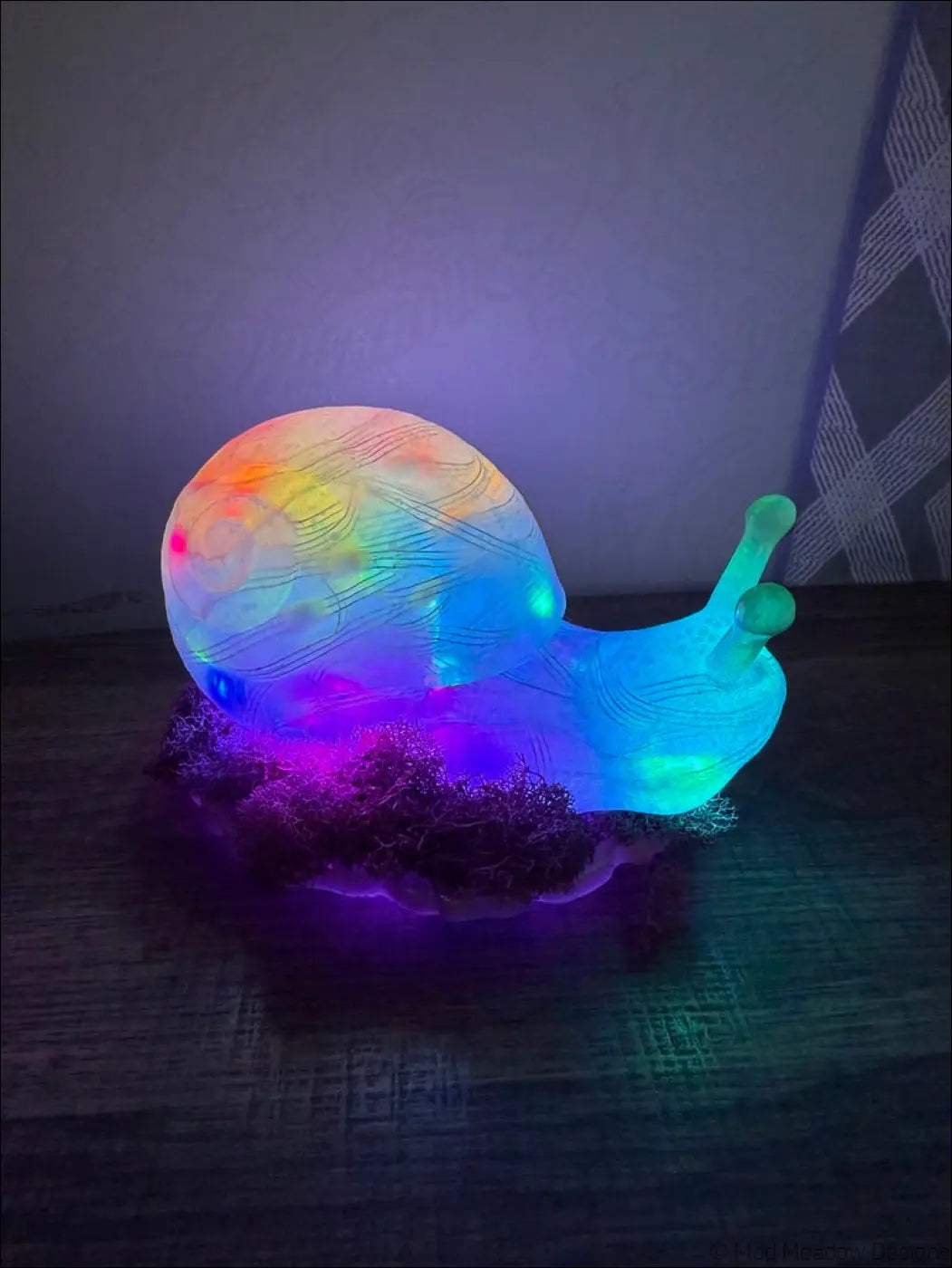 A glowing translucent snail led lamp with a rainbow iridescent shell and blue body on artificial moss light usb app controlled