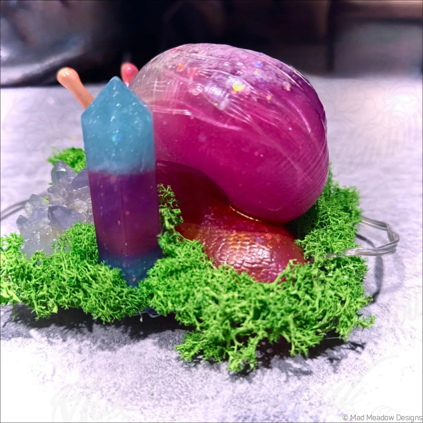 A vibrant translucent pink giant snail figurine with orange antennae resting on moss next to a rgb usb light crystal