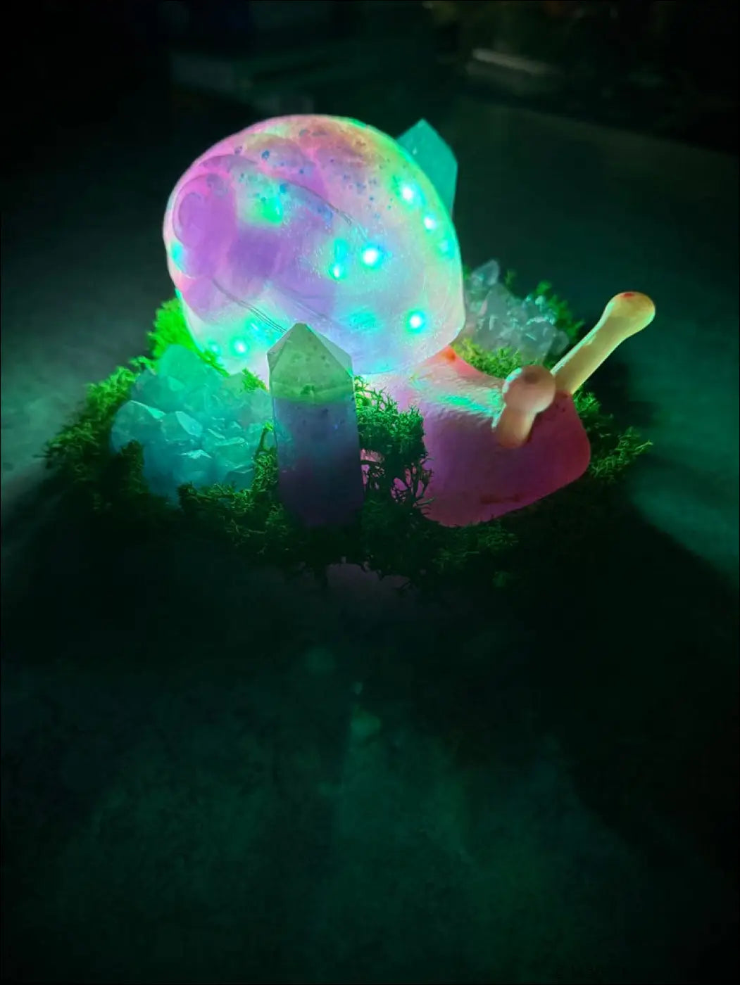 A glowing giant snail figurine with a rainbow shell and pink body sitting on moss with rgb usb or battery light