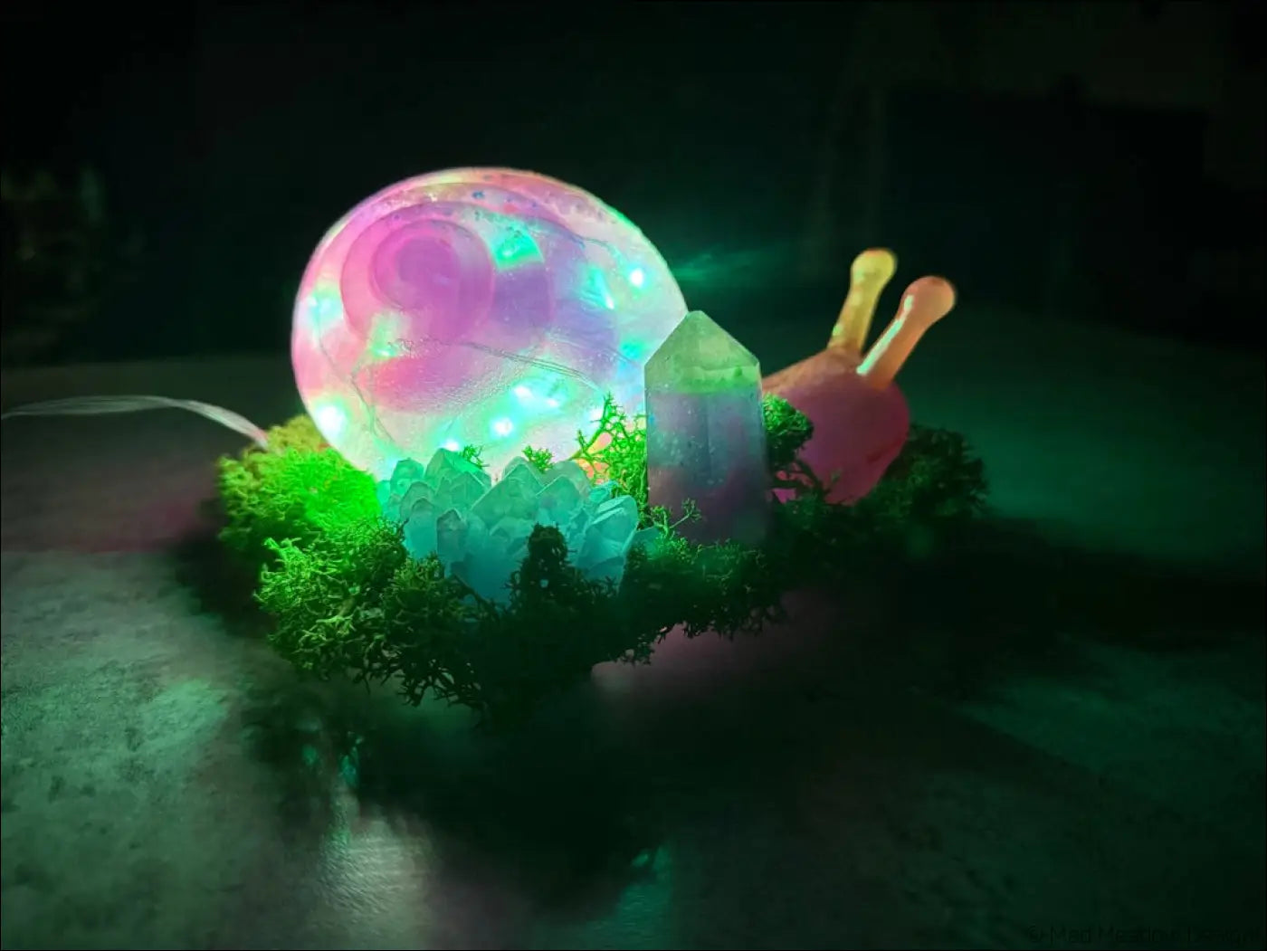 A glowing pink and purple snail-shaped led lamp with a translucent shell on green moss surrounded by crystal accents