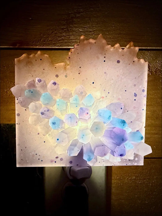 A glowing crystal cluster wall night light with translucent blue and purple crystals on a speckled plug-in base
