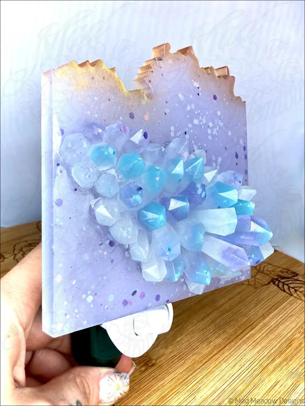 A hand holds a glowing crystal cluster wall night light with blue and white crystals in a lavender epoxy base
