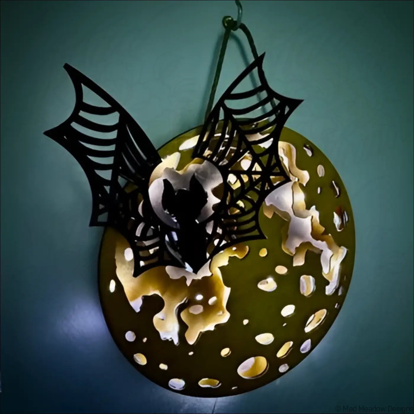 Black bat-shaped laser-cut wood on olive-green plate with white and yellow specks in glowing golden moon hanging light