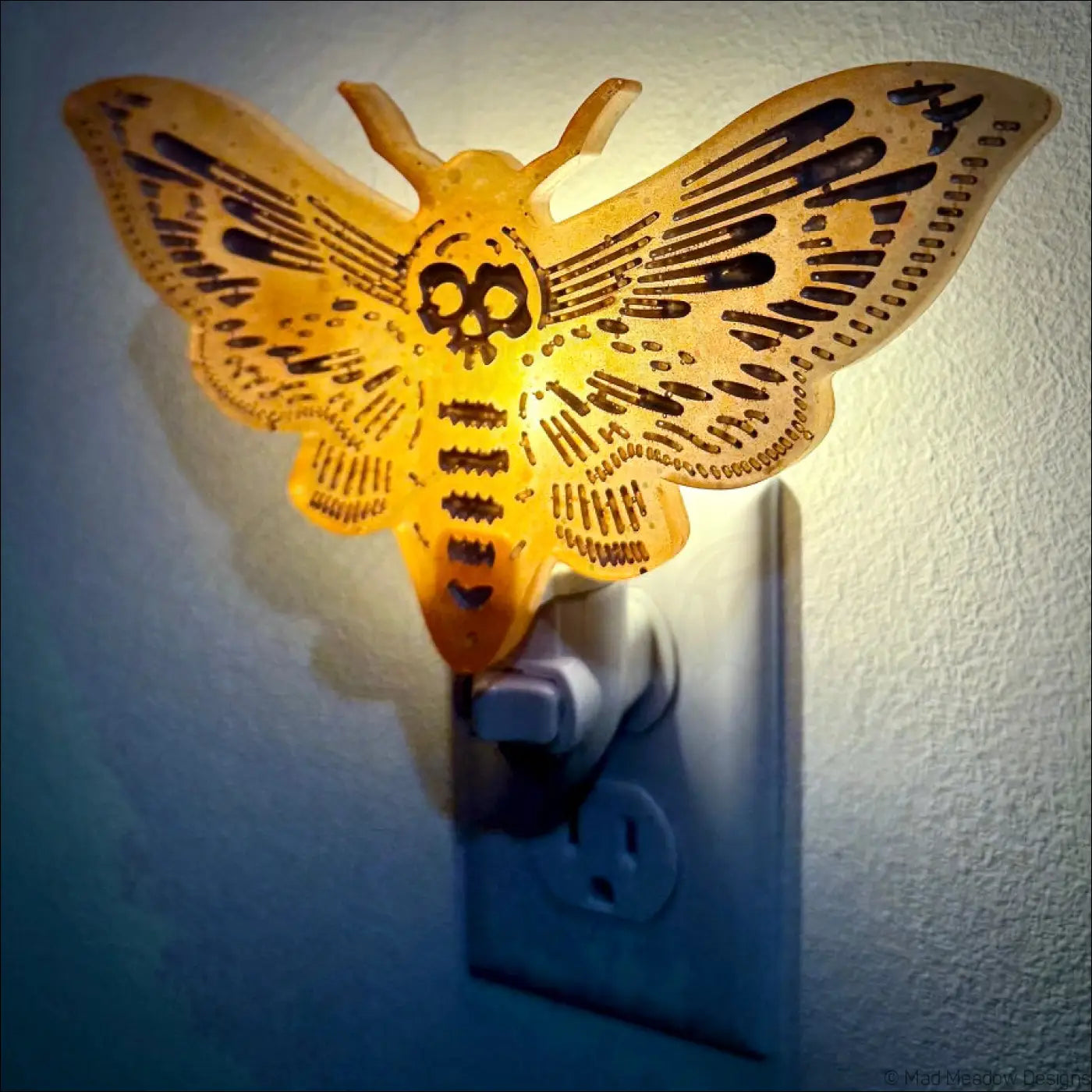Glowing golden death moth resin wall night light with skull design and black cut-out patterns on plug-in base