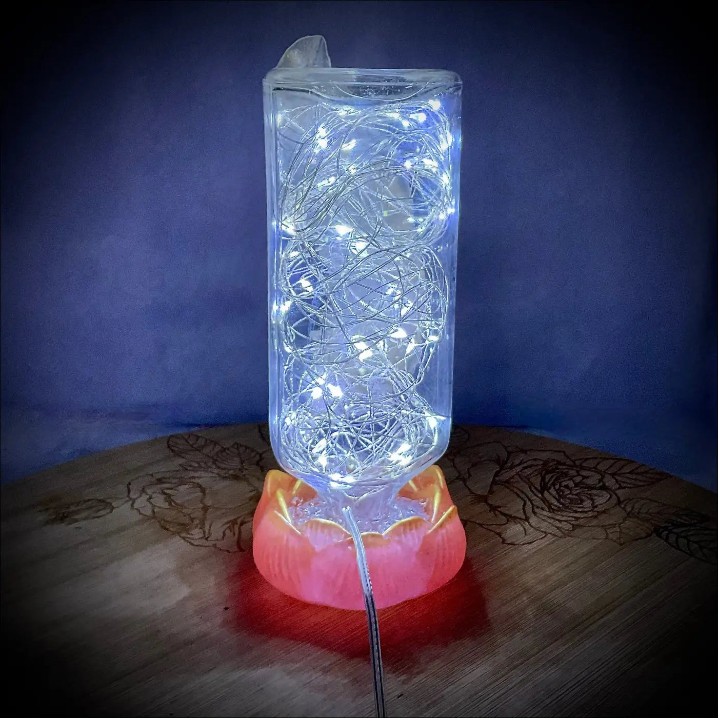 A clear antique glass apothecary bottle lamp with glowing fairy lights on a red-orange lotus flower resin base usb light