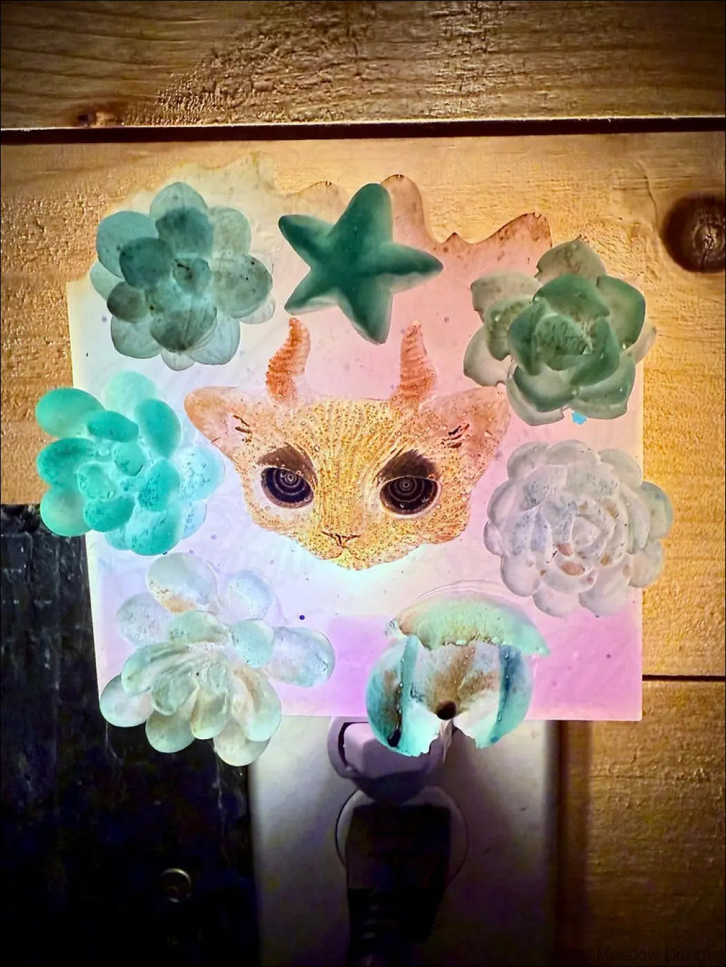 A whimsical horned kitty cat garden wall night light with golden face and green succulents on a soft pink background