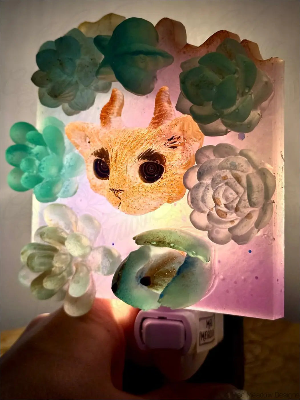 A pink resin horned kitty cat wall night light with an orange cat face and green white succulent designs on plug-in base