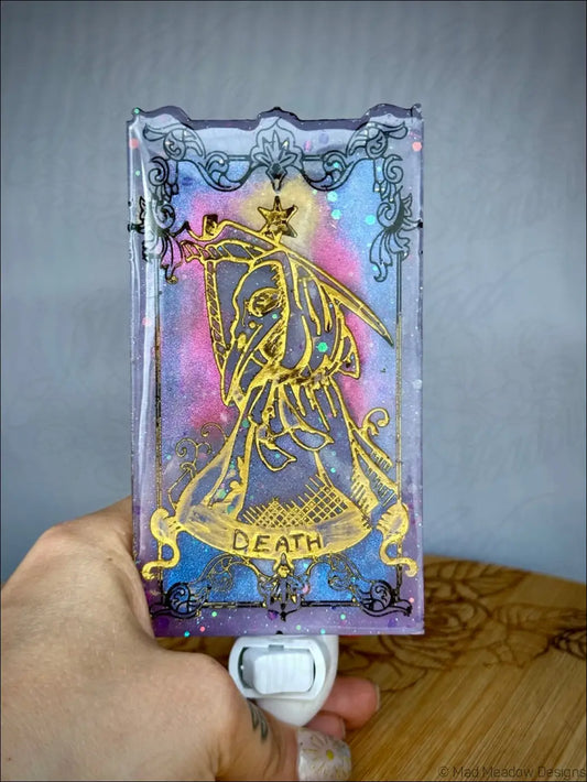 A translucent iridescent death tarot wall night light in pastel purple with gold card design and black glitter filigree accents