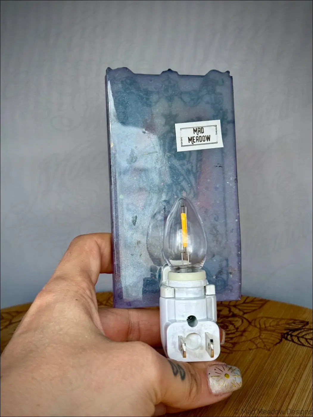 A clear glass iridescent death tarot wall night light with white base held in front of pastel purple mod meadow packaging