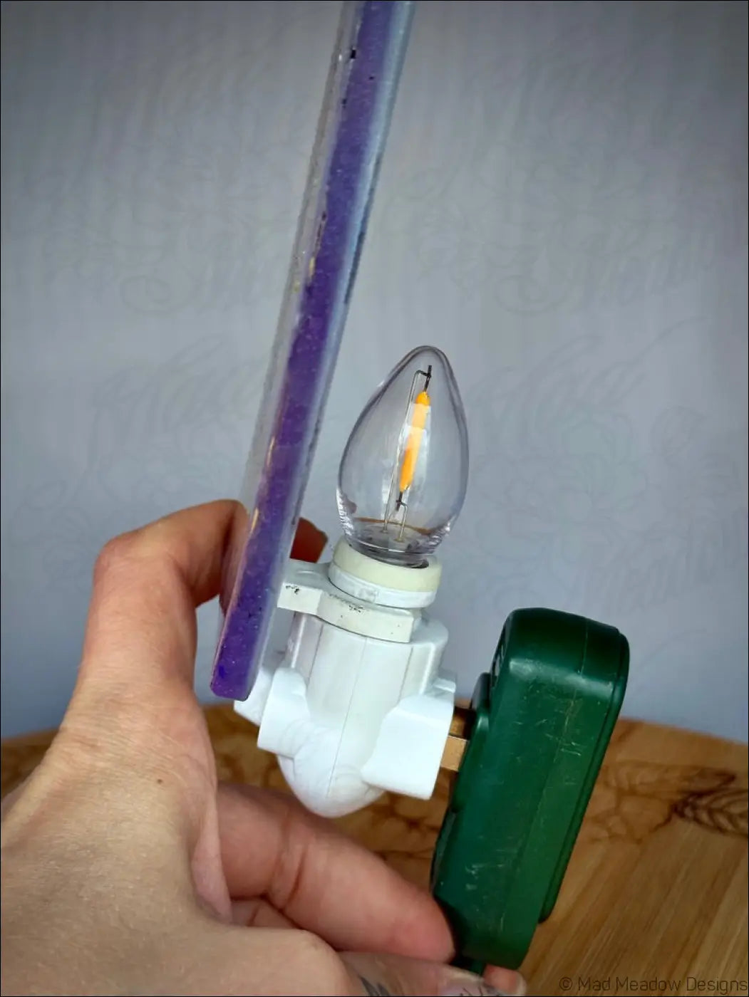 A white plastic plug-in wall night light with an led filament and green adapter held against a pastel purple ruler