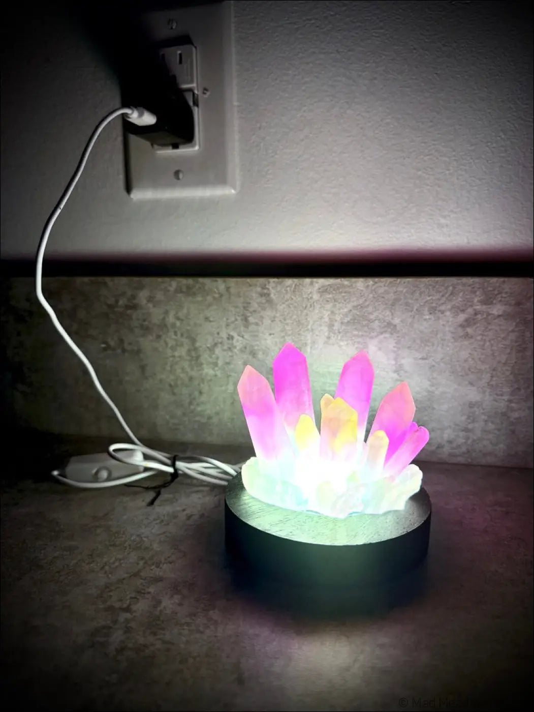 A glowing resin crystal cluster lamp with pink and yellow facets on a white base and dark wooden base stand