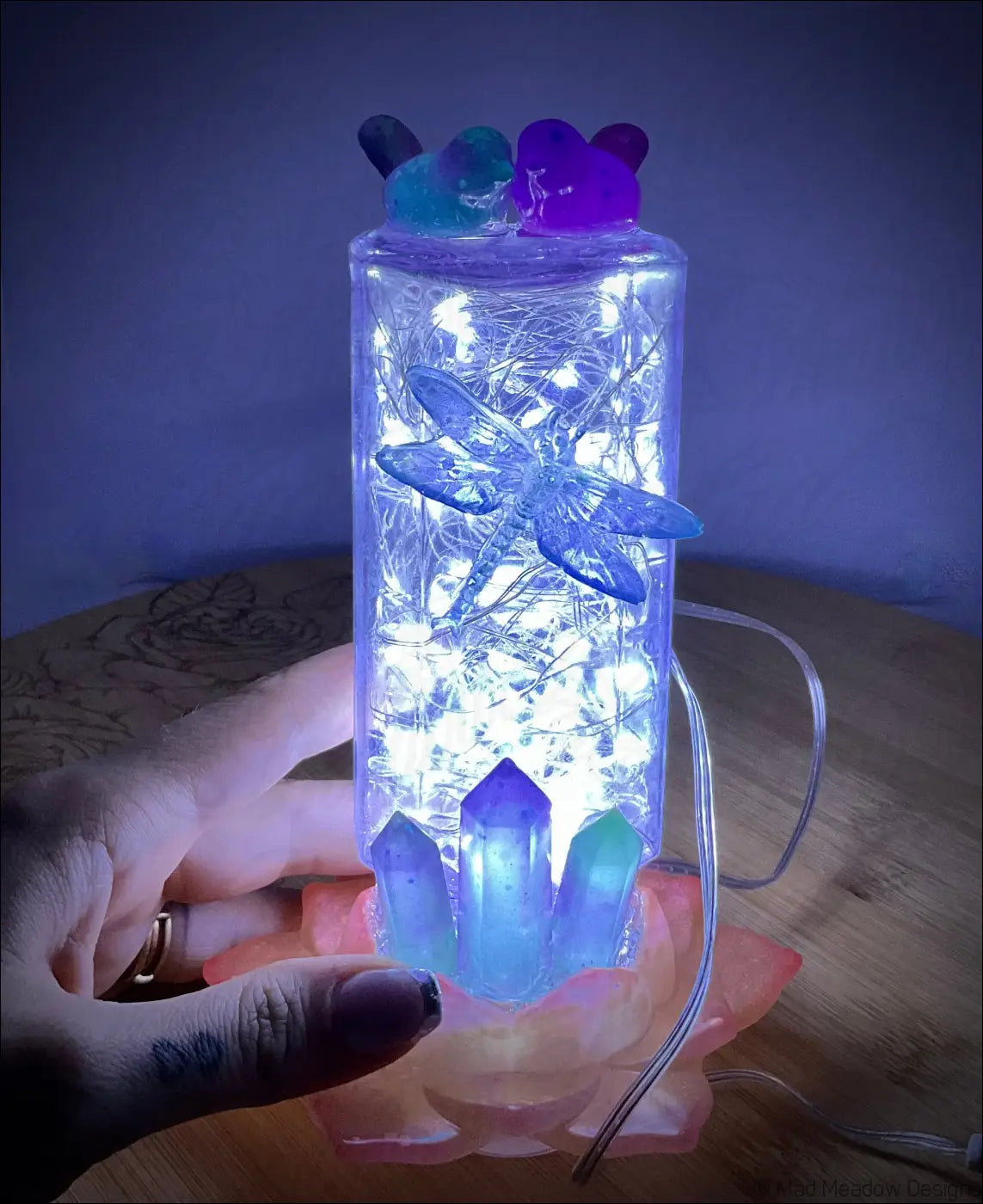 A translucent blue dragonfly resin ornament inside a glowing antique amethyst glass apothecary bottle with crystals and fairy lights