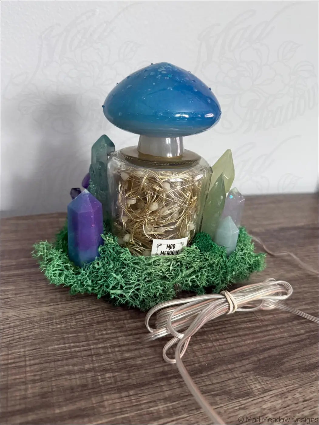Blue mushroom-shaped usb light with clear glass base filled with golden fairy lights surrounded by colorful crystals and moss