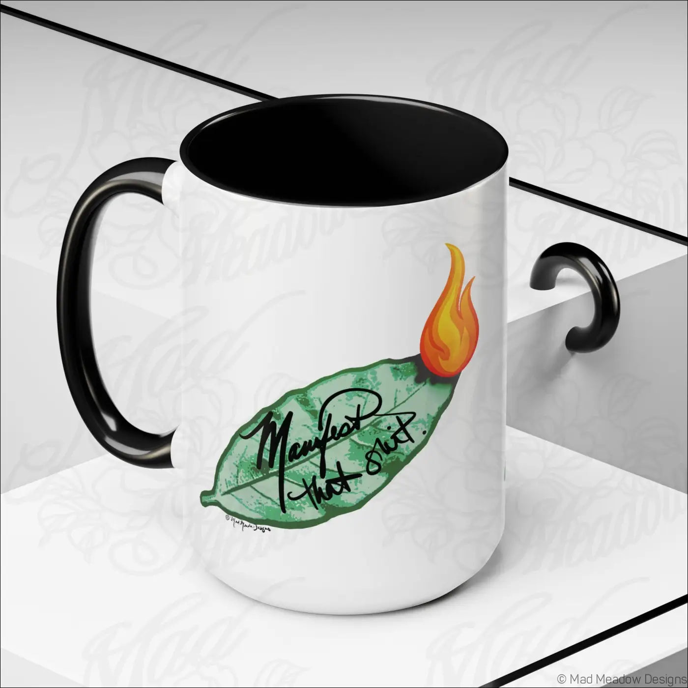 White ceramic coffee mug with glossy finish, black interior and handle, green bay leaf with orange flame and manifest that shit text