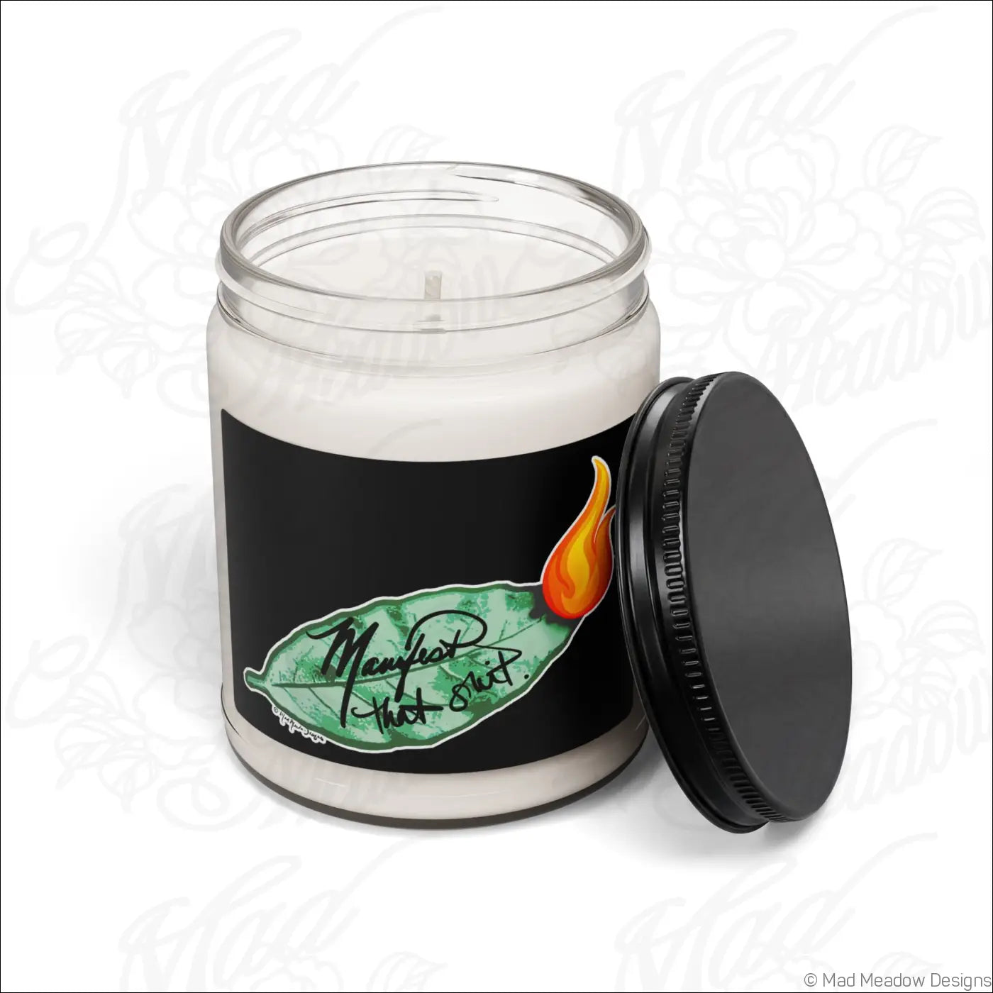 White soy candle in a clear glass jar with black lid and bay leaf label saying manifest that shit with an orange flame
