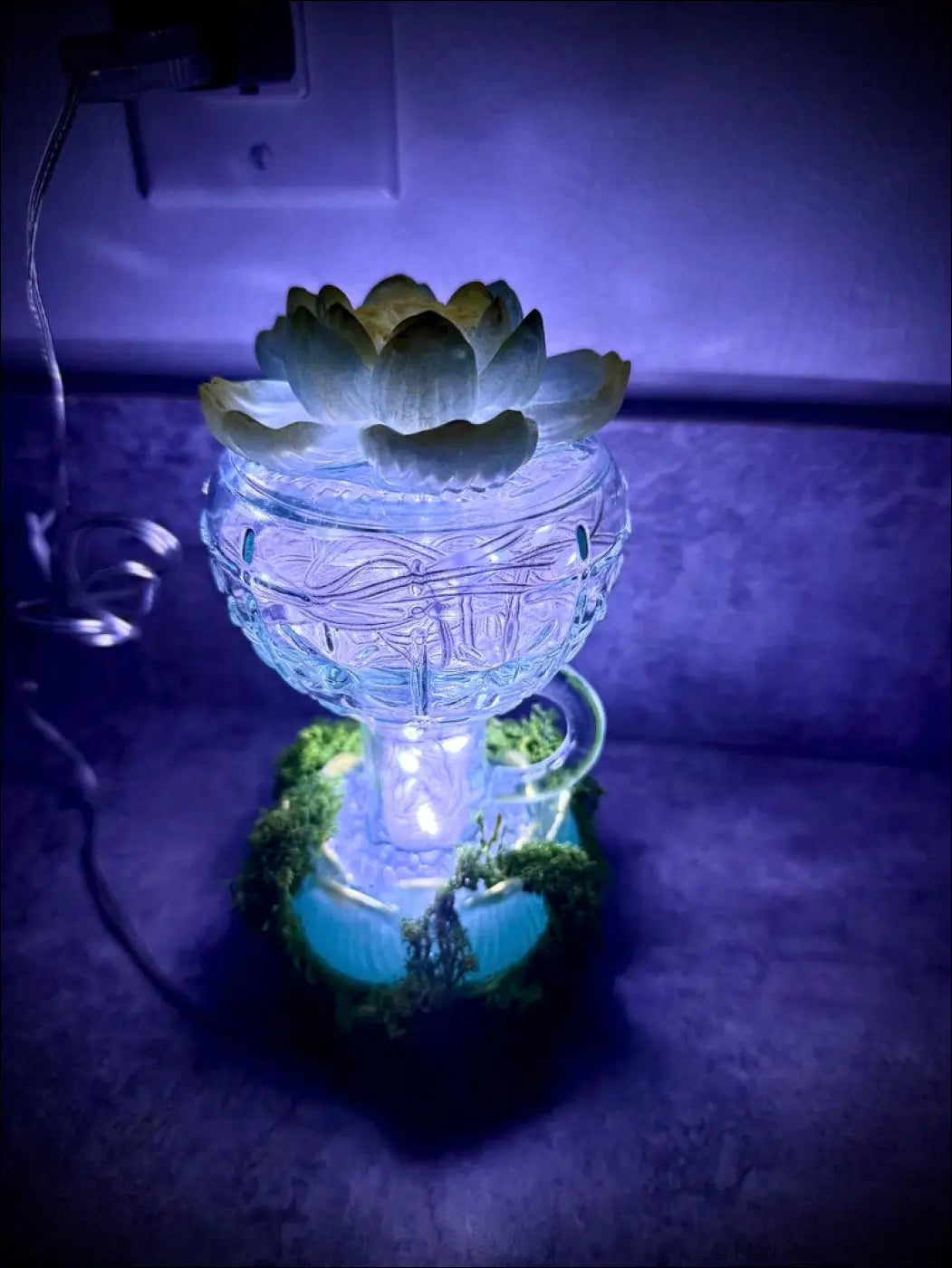 A glowing vintage glass diffuser with mint green and gold lotus design and moss accents with white usb lights