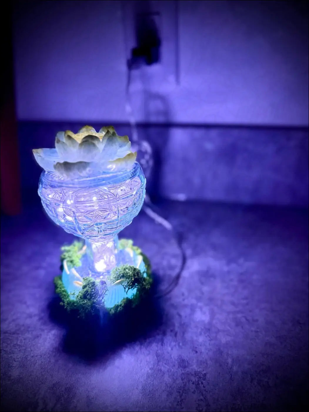 A vintage glass diffuser with mint green and gold lotus details topped with a white lotus and surrounded by green moss