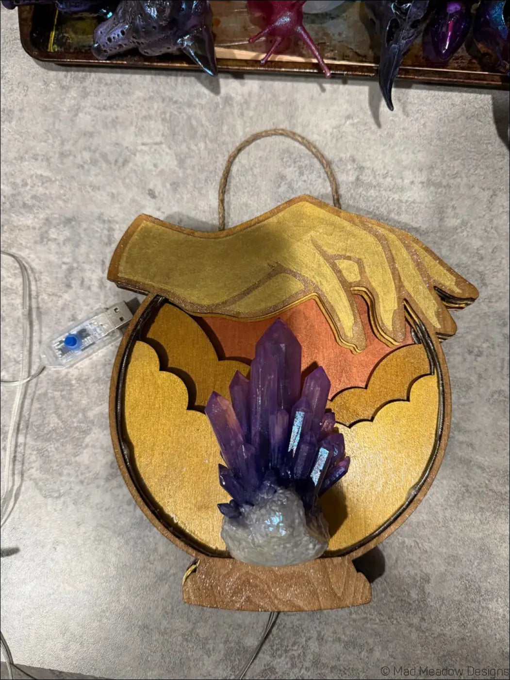 A hand-painted wooden hanging crystal ball with a golden hand over an amethyst cluster and warm orange gold background