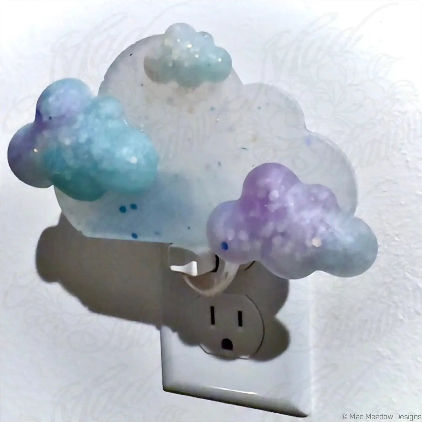 A translucent pastel blue and purple clouds wall night light with glittery resin design on a plug-in base by mad meadow designs