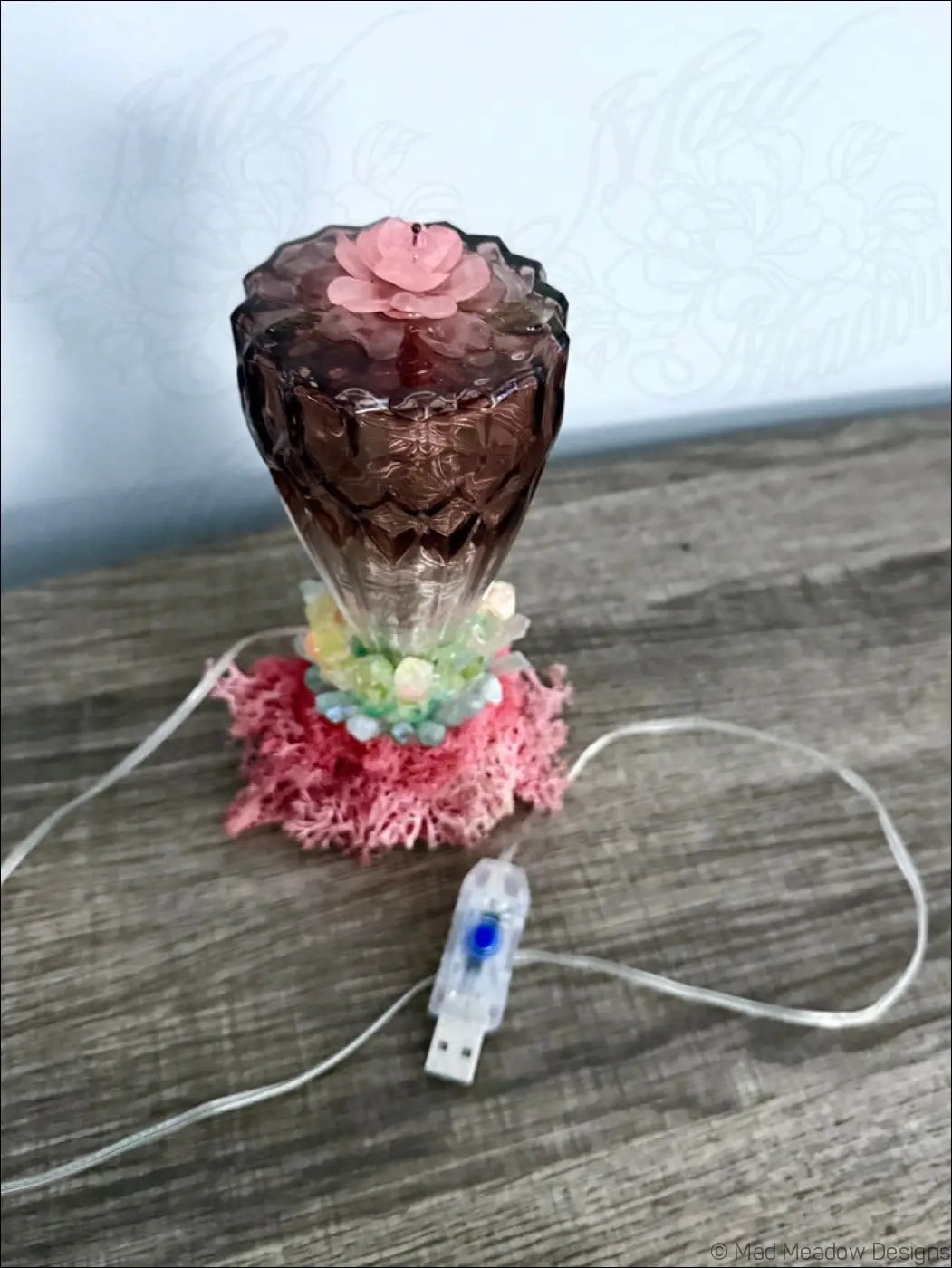 A decorative usb light with dark amber faceted glass body, pink flower top, moss, and quartz crystal points