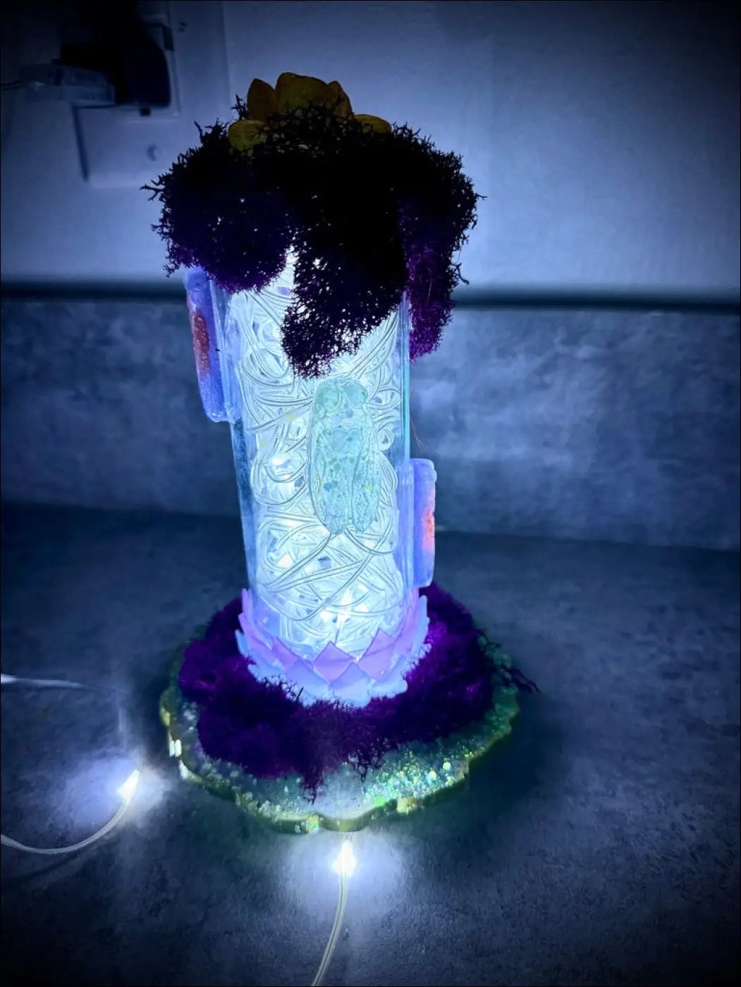 A glowing white usb resin cicadas lamp with intricate etched designs and purple moss accents in a vintage glass bottle