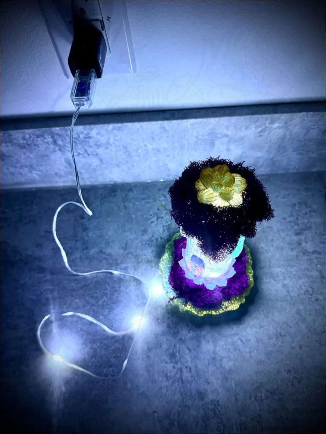 A glowing resin cicadas figurine with a black fuzzy top and white usb light in a vintage glass bottle with plants and moss